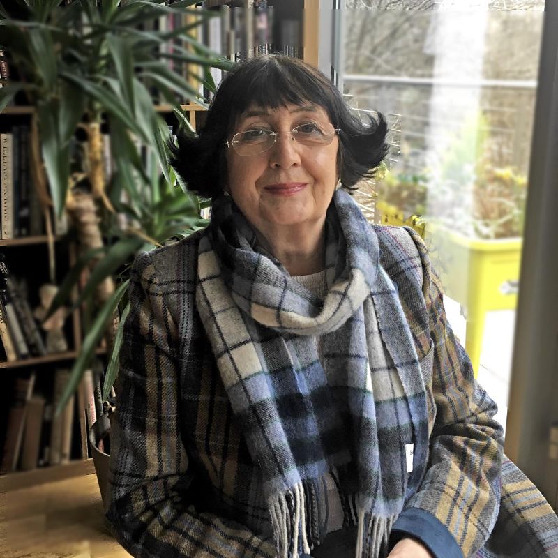 Anita Manning with short black hair and glasses, wearing a plaid scarf and coat, sitting indoors with plants and books in the background.