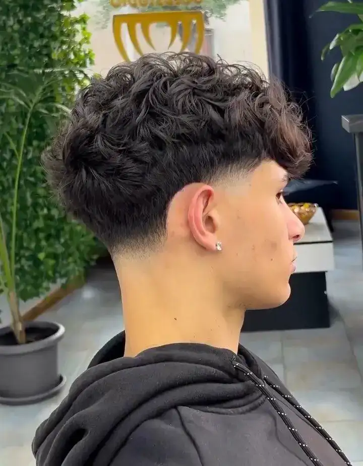 Side profile of a man with a curly fringe and taper fade hairstyle, wearing a black hoodie, in a modern indoor setting.