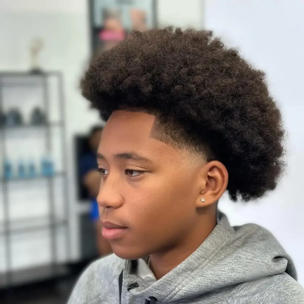 Young person with a low taper fade and curly afro hairstyle wearing a gray hoodie in a salon setting.